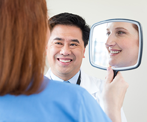 Woman looking in hand mirror with healthcare provider looking on.